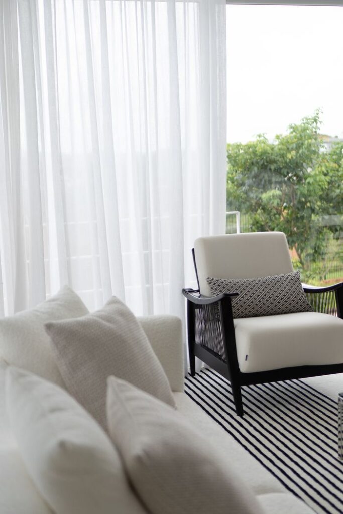 Minimalist white armchair by window with sheer curtains and greenery outside.