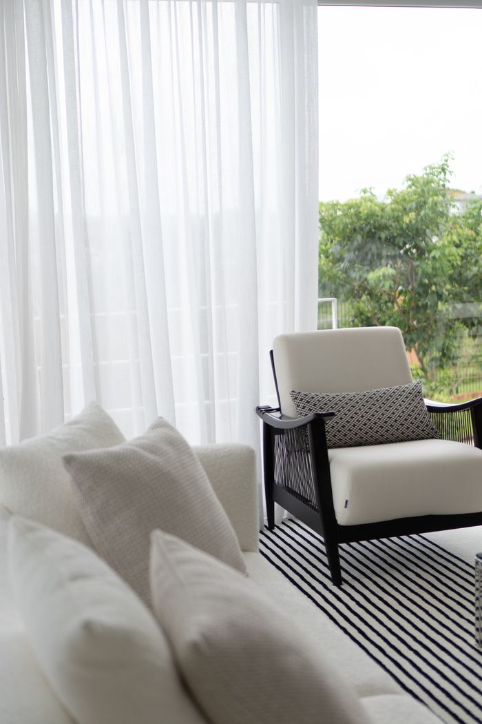 Minimalist white armchair by window with sheer curtains and greenery outside.