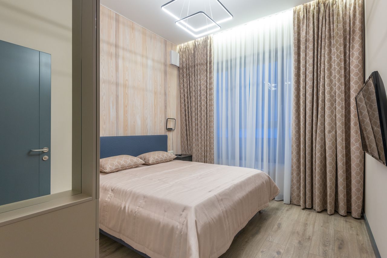 Comfortable bed with cushions and TV hanging on wall in cozy bedroom decorated with curtains